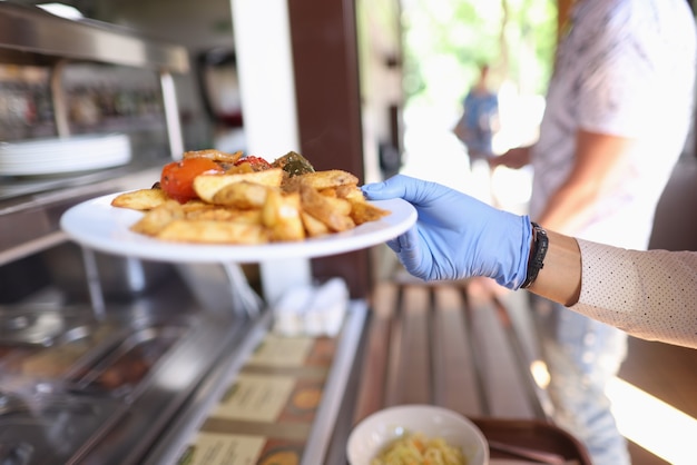 Kobieca ręka w niebieskiej rękawicy medycznej trzyma biały talerz ze smażonymi ziemniakami i duszonymi warzywami