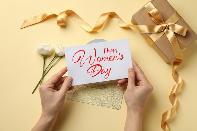 Kobiece ręce trzymają tekst szczęśliwego dnia kobiet na beżowym tle z różami, kopertą i pudełkiem