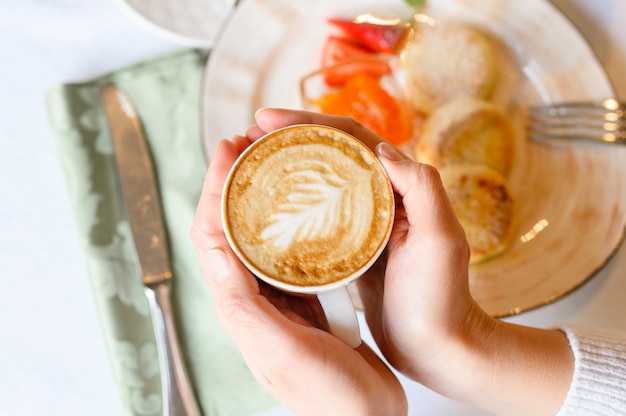 Kobiece ręce trzymając kubek świeżo parzonej kawy z pięknymi liśćmi sztuki latte z pianki na tle stołu z talerzem z jedzeniem. koncepcja porannego śniadania i brunchu