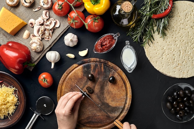 Kobiece ręcznie krojone oliwki na drewnianej desce na kuchennym stole wokół kłamstwa składników do pizzy, warzyw, sera i przypraw