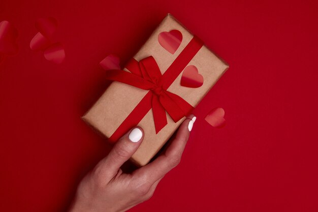 Kobiecej ręki trzymającej Valentine pudełko zawinięte w papier rzemieślniczy z kokardą wstążki i czerwonym sercem