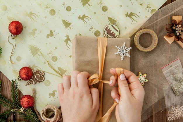 Kobiet ręki zawija Bożenarodzeniowego prezent, zamykają up. Nieprzygotowane prezenty na drewniane elementy dekoracyjne i elementy, widok z góry. Świąteczne lub noworoczne pakowanie DIY.