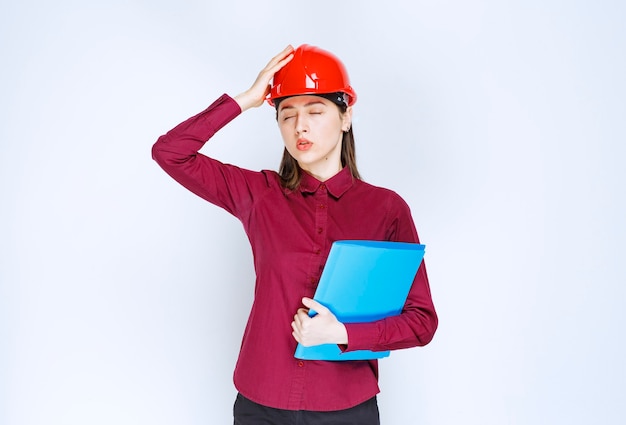 Kobieta architekt w czerwonym kasku trzymając niebieski folder z dokumentami z zmęczonym wyrazem twarzy.
