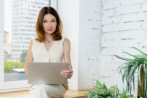 Kobieta biznesu siedzi na parapecie w swoim biurze z laptopem na kolanach Portret kobiety biznesu z rudymi włosami do ramion Miejsce na tekst po prawej stronie