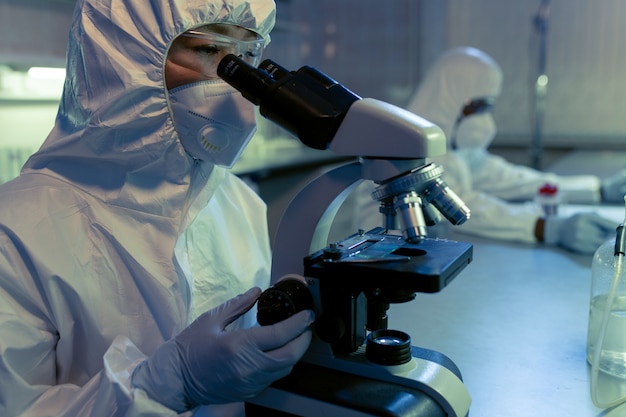 Kobieta chemik w ochronnej odzieży roboczej patrząca przez mikroskop i badająca próbki podczas eksperymentu naukowego w laboratorium