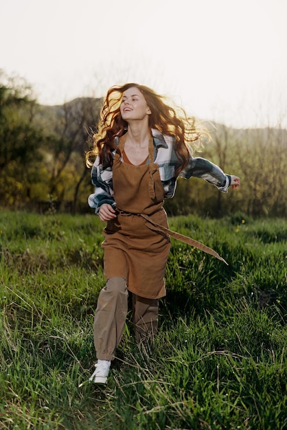 Kobieta chodząca po trawie w przyrodzie uśmiechająca się w rolniczych ubraniach z pięknymi włosami latającymi na wietrze szczęśliwa w promieniach letniego zachodu słońca
