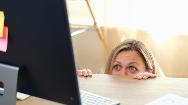 Kobieta chowa się za biurkiem i patrzy na komputerowy błąd w przerażającym filmie w pracy