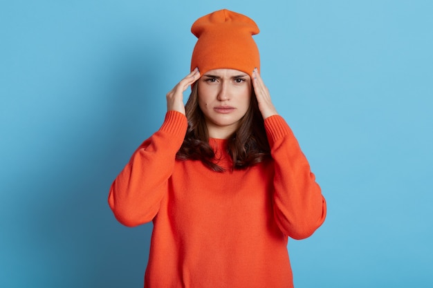 Kobieta cierpiąca na ból głowy, dotykająca skroni obiema rękami, ma migrenę, ma na sobie swobodny pomarańczowy sweter i kapelusz, potrzebuje leczenia, opieki zdrowotnej, pozuje odizolowana na niebieskiej ścianie.