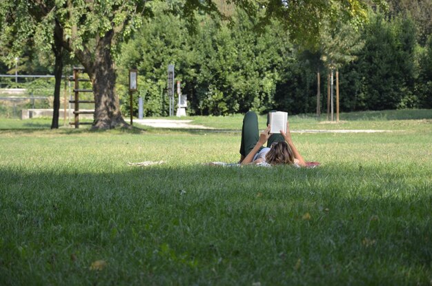 Zdjęcie kobieta czyta książkę leżąc na polu przeciwko drzewom