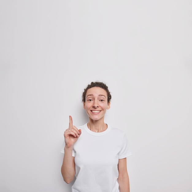 kobieta daje rekomendacje punkty palec wskazujący powyżej na pustej przestrzeni kopii uśmiecha się przyjemnie pokazuje miejsce na kopię ma na sobie luźną koszulkę na białym tle nad białą ścianą