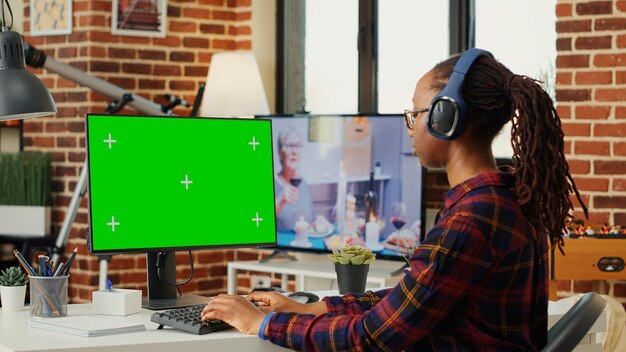 Kobieta freelancer w słuchawkach i korzystająca z zielonego ekranu na pulpicie komputera, siedząca przy biurku i pracująca z izolowanym wyświetlaczem klucza chrominancji. Patrząc na monitor z pustą makietą.
