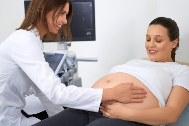 Kobieta ginekolog w fartuchu laboratoryjnym delikatnie dotyka brzucha kobiety w ciąży w nowoczesnej klinice