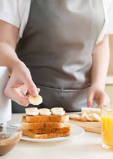 Kobieta gotuje słodkie tosty z masłem orzechowym i bananem. Europejskie śniadanie z tostami i sokiem.
