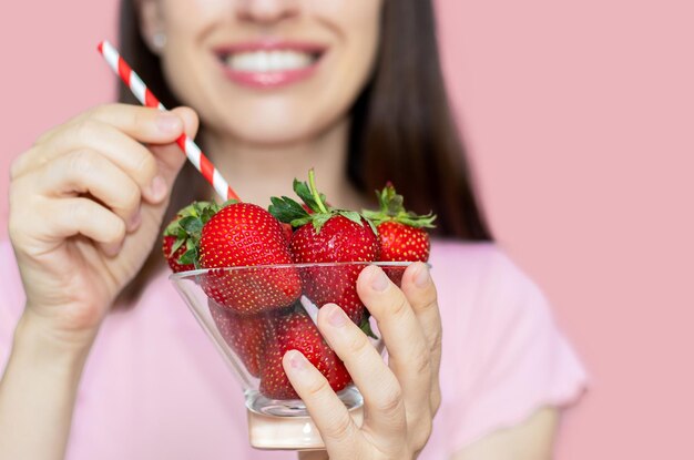 Zdjęcie kobieta gryzie świeże truskawki lub trzyma w ręku szklaną miskę z słomą jak w milkshake witaminy uśmiechając się