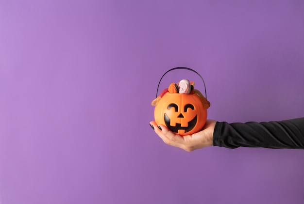 Kobieta Halloweenowa ręka z czarnymi paznokciami trzymająca dynię pełną słodyczy na fioletowym tle z miejscem na kopię