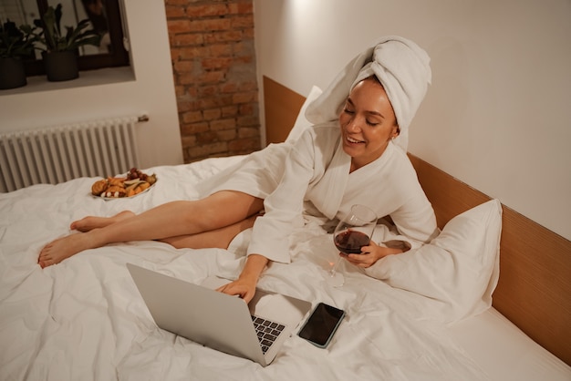Kobieta jest ubrana w szlafrok i biały ręcznik na głowie. Leży na łóżku i surfuje po Internecie przy lampce wina.