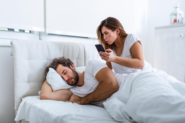 Zdjęcie kobieta jest zazdrosna i podejrzliwa i szpieguje w smartfonie swojego partnera, gdy ten śpi w sypialni. żona szpieguje telefon męża, gdy ten śpi. pojęcie nieufności, zazdrości