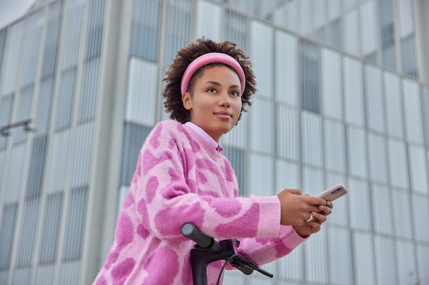 Kobieta Jeździ Na Elektrycznym Skuterze Do Pracy Poza Biurowcami Używa Smartfona Do Wysyłania Smsów Ubrana W Luźny Sweter, Obręcz Na Głowie Skierowana Do Przodu