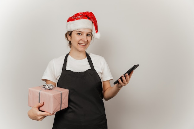 Kobieta kelnerka w santa hat z telefonem i prezentem w dłoniach na szarym tle na białym tle