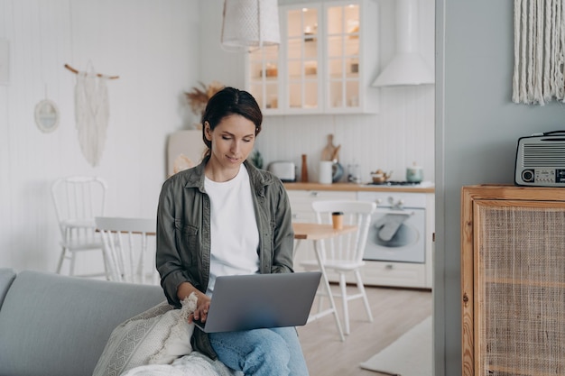 Kobieta korzystająca z laptopa robiąca zakupy lub pracująca na czacie online w sieci społecznościowej, siedząca na kanapie w domu