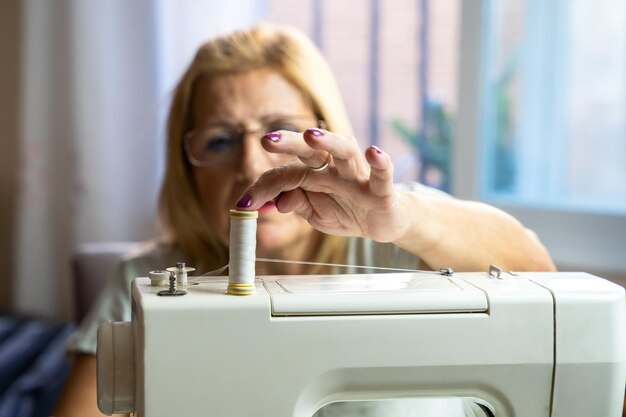 Kobieta krawcowa w okularach siedzi i przygotowuje nić swojej maszyny do szycia