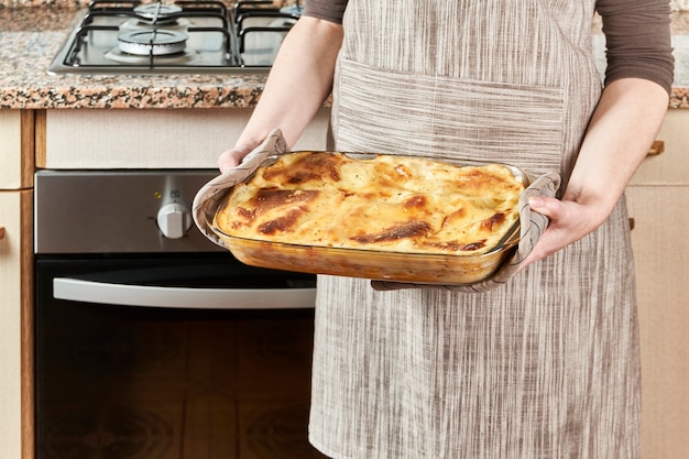 Zdjęcie kobieta kucharka wyjmuje z pieca gorącą lasagne, świeżo upieczoną z roztopionym serem i zapiekaną w swojej kuchni