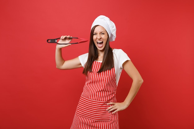 Kobieta kucharz lub piekarz w pasiasty fartuch, biały t-shirt toczek kucharzy kapelusz na białym tle na tle czerwonej ściany. Kobieta trzymać sałatkę kuchenną serwującą plastikową parę szczypiec do grilla. Makieta koncepcja przestrzeni kopii.
