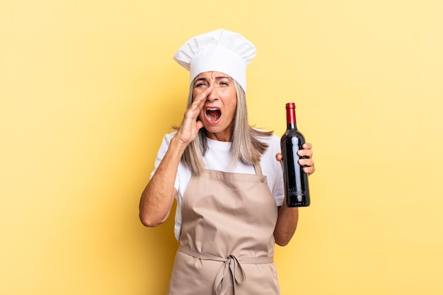 Kobieta kucharz w średnim wieku czuje się szczęśliwa, podekscytowana i pozytywna, wydając wielki okrzyk z rękami przy ustach, wołając trzymając butelkę wina