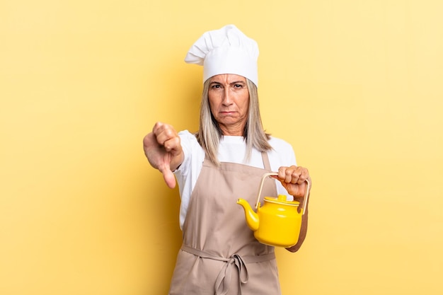 Zdjęcie kobieta kucharz w średnim wieku czuje się zła, zła, zirytowana, rozczarowana lub niezadowolona, pokazując kciuk w dół z poważnym spojrzeniem i trzymając czajnik