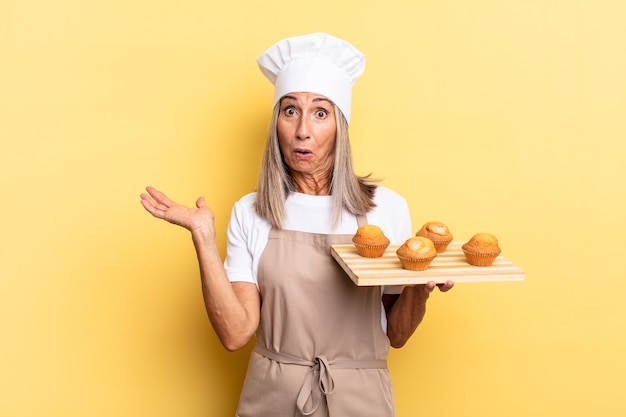 kobieta kucharz w średnim wieku wyglądająca na zaskoczoną i zszokowaną, z opuszczoną szczęką, trzymająca przedmiot z otwartą dłonią z boku i trzymającą tacę z babeczkami