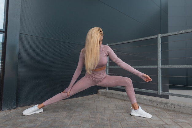 Kobieta ładna blond fitness model z długimi włosami w modnej odzieży sportowej z białymi trampkami robi rozciąganie przed joggingiem i rozgrzewką w mieście