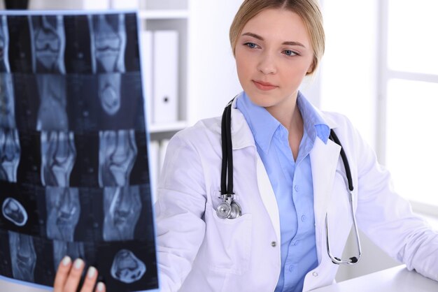 Kobieta lekarz bada zdjęcie rentgenowskie w pobliżu okna w szpitalu. Chirurg lub ortopeda w pracy. Koncepcja medycyny i opieki zdrowotnej. Niebieska bluzka terapeuty wygląda dobrze.