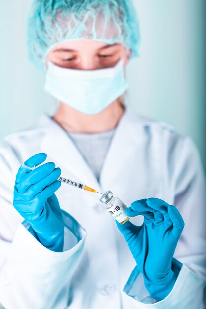Kobieta lekarz lub pielęgniarka w mundurze i rękawiczkach noszących maskę ochronną w laboratorium trzymająca fiolkę z lekarstwem butelka szczepionki z etykietą szczepionki przeciwko koronowirusowi COVID-19