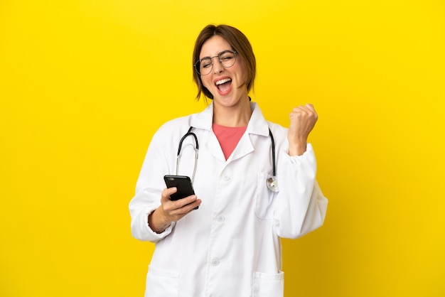 Kobieta lekarz na białym tle na żółtym tle z telefonem w pozycji zwycięstwa