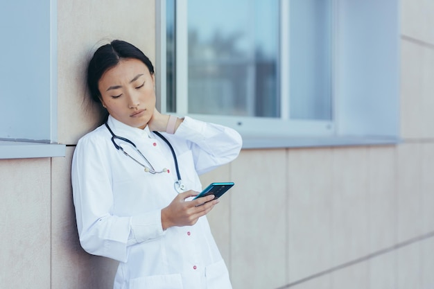 Kobieta-lekarz-pielęgniarka ma silny ból szyi, nieszczęśliwa Azjatka w pobliżu przychodni, używa telefonu, czyta z aplikacji medycznej