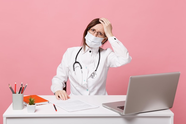 Kobieta lekarz siedzi przy biurku na komputerze z dokumentem medycznym w masce na twarz w szpitalu na tle pastelowej różowej ściany. Kobieta w okularach medycznych suknia stetoskop Koncepcja medycyny opieki zdrowotnej