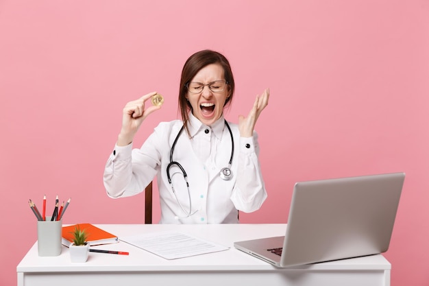Kobieta lekarz siedzi przy biurku pracuje na komputerze z dokumentem medycznym trzymaj monetę w szpitalu na białym tle na pastelowym różowym tle ściany. Kobieta w medycznym stetoskopie toga okularów. Koncepcja medycyny opieki zdrowotnej