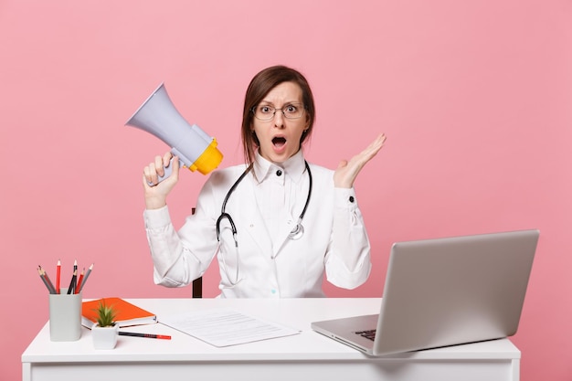 Zdjęcie kobieta lekarz siedzi przy biurku przy pracy na komputerze z dokumentem medycznym trzymaj megafon w szpitalu na białym tle na pastelowym różowym tle. kobieta w medycznym stetoskopie toga okularów. koncepcja medycyny opieki zdrowotnej.
