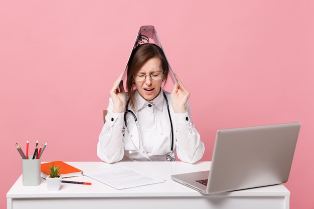 Kobieta lekarz siedzi przy biurku w pracy na komputerze z folderem dokumentów medycznych w szpitalu na białym tle na pastelowym różowym tle ściany. Kobieta w medycznym stetoskopie toga okularów. Koncepcja medycyny opieki zdrowotnej