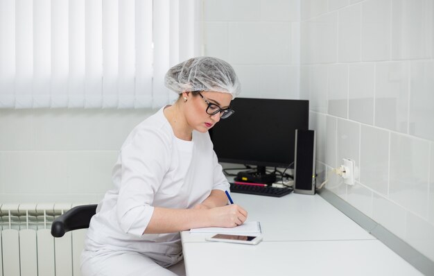 Kobieta lekarz w okularach i białym garniturze medycznym siedzi przy biurku, robiąc notatki w zeszycie w gabinecie kliniki