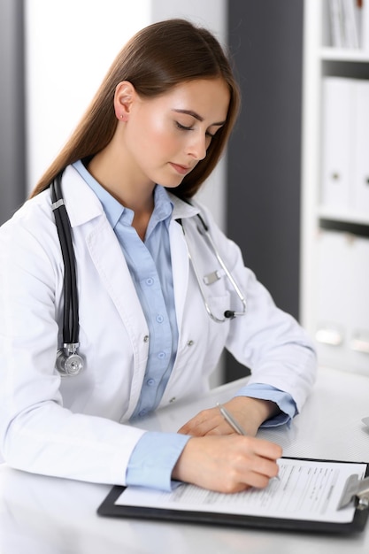 Kobieta lekarz wypełniania formularza medycznego siedząc przy biurku w biurze szpitala. Lekarz w pracy. Koncepcja medycyny i opieki zdrowotnej.