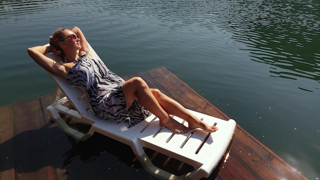 Kobieta leży na leżaku w okularach przeciwsłonecznych i jedwabnym szalu boho Dziewczyna odpoczywa na zalanym drewnem podwodnym molo Bruk jest zalany wodą w jeziorze