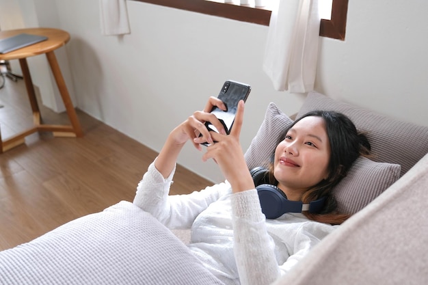 Kobieta leży oparta na wygodnej sofie i używa swojego nowoczesnego telefonu komórkowego do czatowania z przyjaciółmi, przewijaj w aplikacji zakupowej lub w mediach społecznościowych
