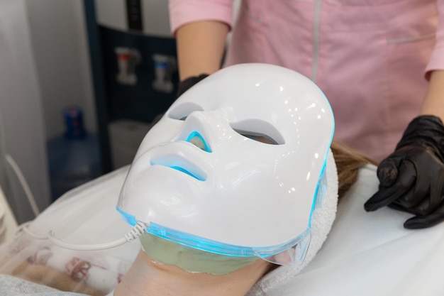 Kobieta leży w łóżku z maską na twarz do terapii światłem i relaksuje się. Maska świetlna led do pielęgnacji skóry twarzy, zabieg kosmetologii profesjonalnej.