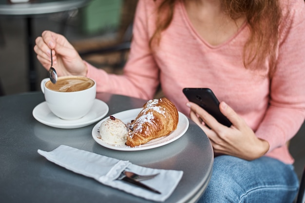 Kobieta ma śniadanie w caffe i przy użyciu smartfona. Dziewczyna na czacie i za pomocą Internetu z telefonem podczas przerwy na kawę z rogalikiem