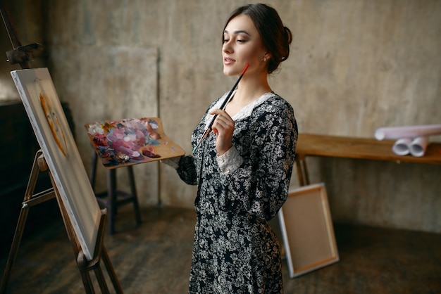 Kobieta malarz z pędzla i palety pozuje w studio sztuki. Artystka w miejscu pracy, mistrz kreatywności przy sztalugach w pracowni