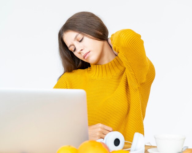 Kobieta masuje ból szyi od długiego czasu pracy przy komputerze. Piękna młoda dama w jasnożółtym swetrze siedzi przy biurku