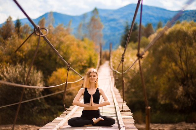 Kobieta medytująca w pozycji lotosu na moście nad górską rzeką w jesiennym lesie