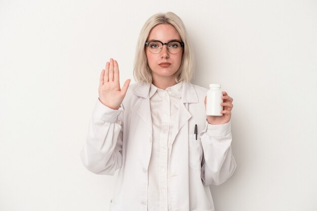 Kobieta młody farmaceuta trzymając pigułki na białym tle stojący z wyciągniętą ręką pokazując znak stop, uniemożliwiając.