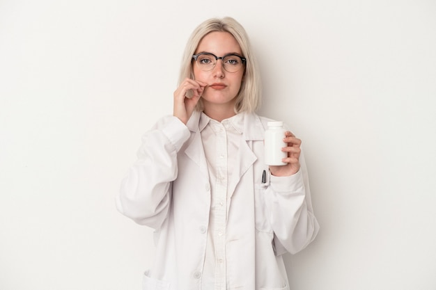 Kobieta młody farmaceuta trzymając pigułki na białym tle z palcami na ustach zachowując tajemnicę.